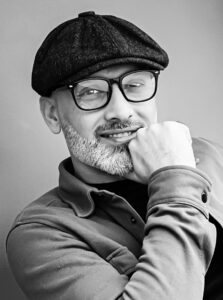 Smiling man wearing glasses and a flat cap at Kobani Festival in black and white.